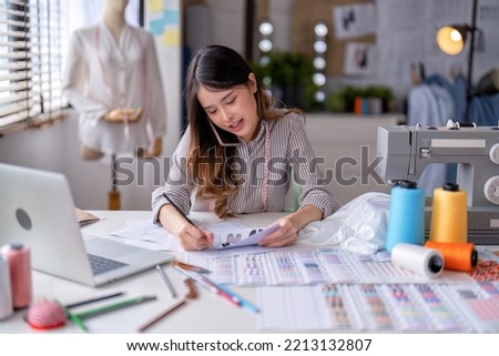 Young professional clothes fashion textile creative thinking designer sitting near sewing machine use mobile phone to discuss talking with customer in studio office room