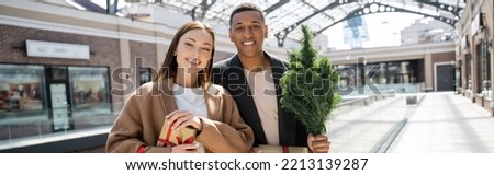 trendy interracial couple with gift boxes and small christmas tree smiling near blurred mall, banner