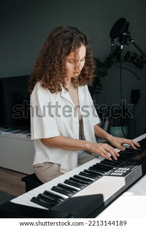 Young attractive woman with curly hair playing music on synthesizer at home. Midi keyboard.