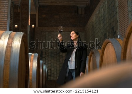 Woman in the wine cellar with barrels in background drinking and tasting wine. Royalty-Free Stock Photo #2213154121
