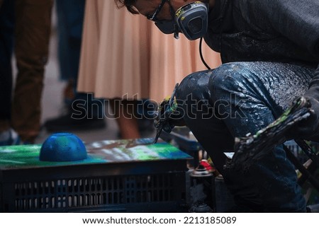 a man dressed in protective clothing on the street creates a spray art picture with spray paint cans, he was surrounded by a crowd of people and looks