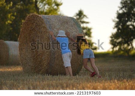 A boy and a girl merrily roll a reel of hay across a mowed field in the summer