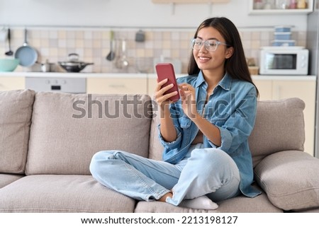 Young asian woman in eyeglasses calling friend online using mobile phone sitting on sofa at home. Happy millennial teen girl checking social media, playing game or ordering delivery holding smartphone Royalty-Free Stock Photo #2213198127