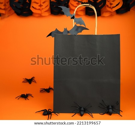 Halloween shopping and sale concept. Paper black bag and decor for Halloween on orange background. Mock up. Copy space. Selective focus.