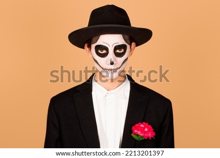 Portrait of spooky handsome guy made makeup for Halloween event, has image of ghost, red rose flower in pocket of white shirt, wears black hat, has scary look, dressed in zombie attire