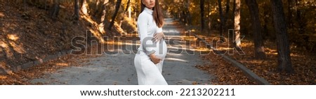 Portrait of young pregnant pretty woman with hands on belly, wearing white dress on background of golden autumn park. Panoramic banner view.