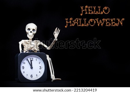 A human skeleton waving his hand, greeting holding an black alarm clock on a black background with the text Hello Halloween, copy space. It's Halloween time. Greeting card