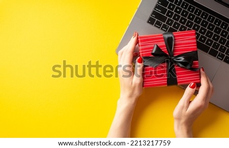 Cyber monday concept. First person top view photo of female hands holding red giftbox with ribbon bow over laptop on isolated yellow background with empty space