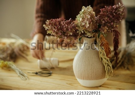 Dried hydrangea flowers in vase on background of woman arranging dried grass in wreath on wooden table. Making stylish autumn wreath on rustic table. Fall decor and arrangement in farmhouse Royalty-Free Stock Photo #2213228921