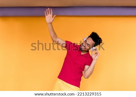 Exited young dark-skin guy with happy emotions closed eyes, holding up hand and dancing while listening music and having fun over yellow background .
