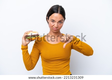 Young hispanic woman holding a burger isolated on white background showing thumb down with negative expression