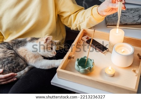 Mental health, mindfulness, wellbeing. Faceless portrait of relaxed young woman with cat sitting near table with burning candles and aroma sticks at home and do meditation and relaxation