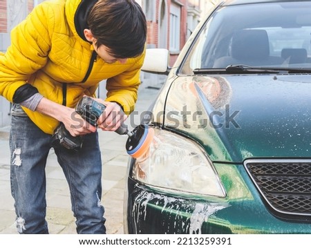A young caucasian guy in a yellow jacket is drilling a drill with a disk with a sponge and polishing the headlight of his car with detergent soap on the city street in front of the house, close-up sid