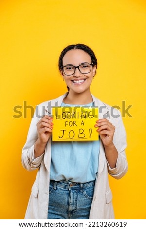 Vertical photo of positive successful smart young woman, creative specialist looking for a job, stands on isolated orange background, shows a sign looking for a job, looks at camera, smile