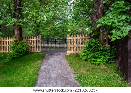 The opening of a wooden picket fence at the end of a concrete pathway. There are large mature lush green maple trees with sprigs sticking out at the bottom of the tree trunks covered in leaves.  Royalty-Free Stock Photo #2213263237