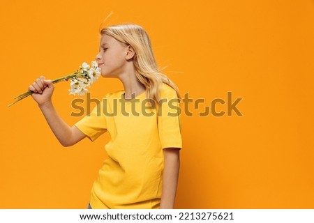 a beautiful, cute girl of school age stands with a bouquet of daisies in her hands on an orange background and smells the flowers. Studio photography with empty space for inserting advertising text