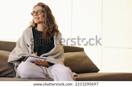 Journaling and self-awareness. Mature woman looking away thoughtfully while writing in her journal at home. Senior woman writing down her thoughts while sitting on a couch. Royalty-Free Stock Photo #2213290697