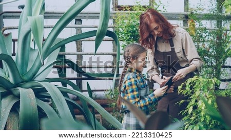 Young female farmer is teaching her cheerful daughter to work with plants, they are touching green plant, talking and laughing. Farming, childhood and family concept.