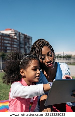 African woman sitting in the grass with her young daughter and using tablet. They are enjoying a sunny day.