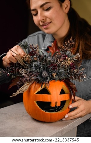 woman florist gently decorate lovely autumn arrangement of thistles flowers and plants in bright orange pumpkin with carved eyes and mouth. Beautiful floral decor for halloween. Close-up Royalty-Free Stock Photo #2213337545