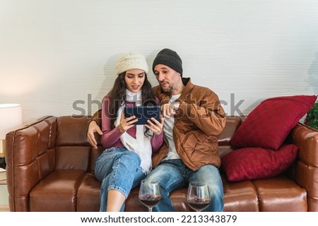 European couple man and woman smiling together looking pointing at tablet mobile device sitting on a brown leather sofa in front of white wall. Male and female wearing warm winter clothing.