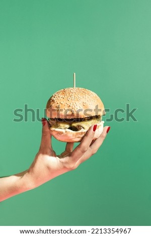 Modern scene with womans hand holding a burger against green background. Minimal composition with copy space. Fast food and street food concept.