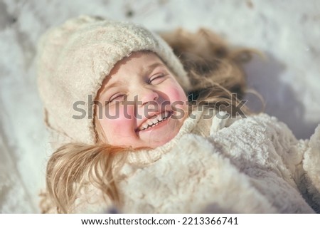 Beautiful little girl lies on the snow and laughs at the camera close-up. A girl in a white coat laughs in the snow. Happy childhood in winter. Games in the snow.