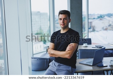 Portrait of a smiling young man in a black t-shirt against the background of a modern office. Stylish guy in casual clothes
