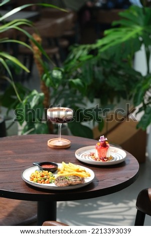 Steak on plate, a plate of panna cotta, and drink in tall glass. Food and drink on the table. Plant in pot as background. Steak, panacota dan minuman di meja. Royalty-Free Stock Photo #2213393039