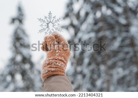 hand in mitten holding decorative transparent snowflake, winter forest natural background. Christmas and New Year holidays concept. symbol of winter festive season.