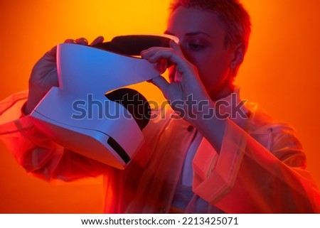 Crop young futuristic woman with short blond hair in transparent raincoat wearing modern VR headset against orange background in neon studio