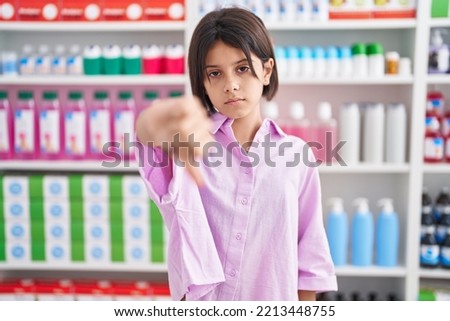Young girl at pharmacy drugstore looking unhappy and angry showing rejection and negative with thumbs down gesture. bad expression. 