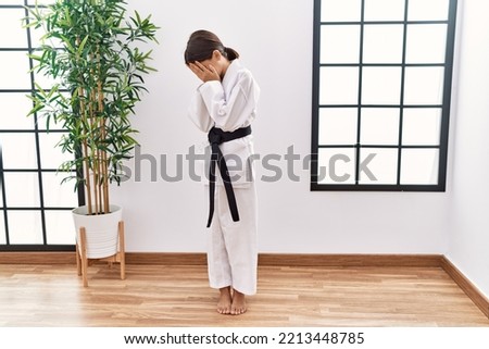 Young hispanic girl wearing karate kimono and black belt with sad expression covering face with hands while crying. depression concept. 