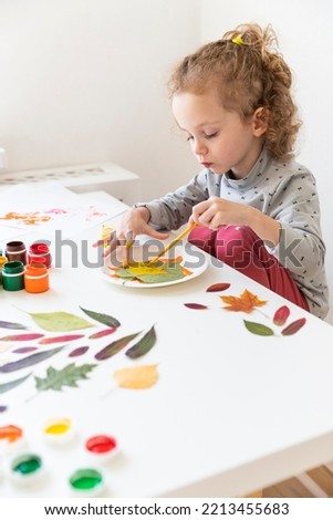 A little girl makes crafts on an autumn theme. A three-year-old girl paints a sheet with yellow paint with her left hand. The girl got her fingers dirty. There is a white wall in the background
