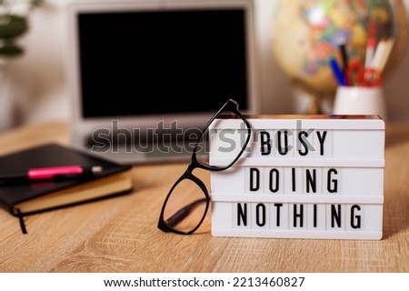 Busy doing Nothing. light box sign sitting on top of a desk. glasses and a desktop. in the background is a computer and a glass with writing utensils. High quality photo