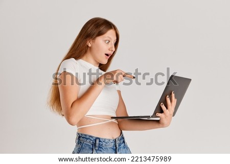 Portrait of young girl looking and pointing at laptop screen isolated over grey background. Information and education. Concept of emotions, lifestyle, facial expression, beauty. technology