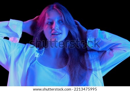 Portrait of young beautiful girl posing in white casual clothes isolated over black background in blue neon light. Delightful. Concept of emotions, lifestyle, facial expression, beauty, fashion, youth