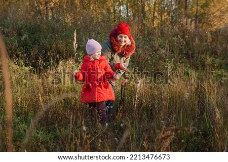 A little girl in a red coat walks in nature in an autumn grove with her grandmother. The time of the year is autumn.