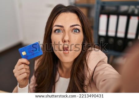 Young brunette woman working at small business ecommerce holding credit card making fish face with mouth and squinting eyes, crazy and comical. 