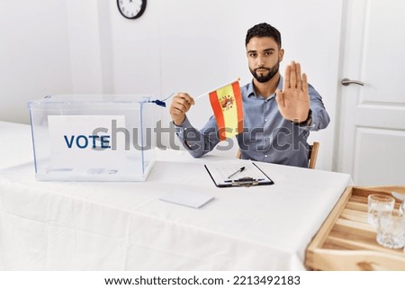 Young handsome man with beard at political campaign election holding spain flag with open hand doing stop sign with serious and confident expression, defense gesture 