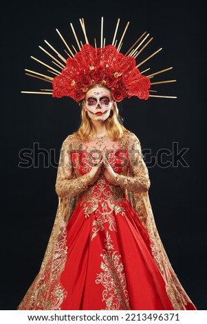 Beautiful Santa Muerte in a festive golden-red dress and floral headdress stands on a black background. Portrait of Calavera Catrina. Dia de los Muertos. Sugar skull girl. Day of The Dead. Halloween.