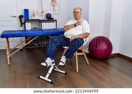 Senior caucasian man at physiotherapy clinic using pedal exerciser cheerful with a smile of face pointing with hand and finger up to the side with happy and natural expression on face 