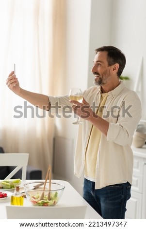 cheerful man with glass of white wine taking selfie near fresh vegetables and salad in kitchen