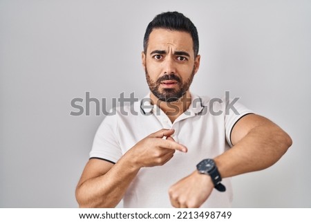 Young hispanic man with beard wearing casual clothes over white background in hurry pointing to watch time, impatience, upset and angry for deadline delay  Royalty-Free Stock Photo #2213498749