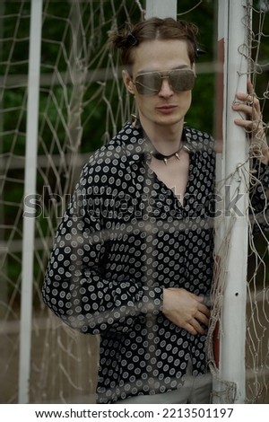 Teenage guy on the football field. Rope mesh around. Trees and sand in the background. Strange clothes. Eclectic style. Emotional. Cool glasses. Male model with a beautiful face. European style.