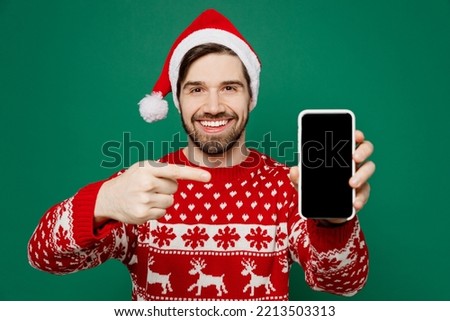 Merry young man wear red knitted sweater Santa hat posing use point finger on blank screen mobile cell phone isolated on plain dark green background studio. New Year 2023 holiday celebration concept