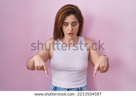 Brunette woman standing over pink background pointing down with fingers showing advertisement, surprised face and open mouth 