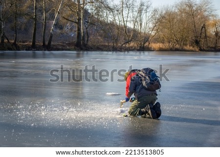 A man in a winter jacket, trousers and with a backpack and spinning nearbue diligently pierces the ice on the frozen river with an axe. Back view Royalty-Free Stock Photo #2213513805