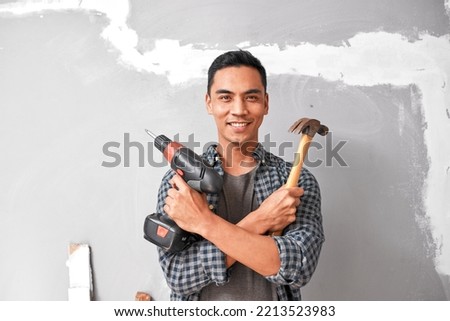 A young Asian man smiles while holding drill and hammer for home DIY improvement Royalty-Free Stock Photo #2213523983