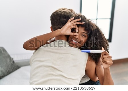 Young interracial couple holding pregnancy test result smiling happy doing ok sign with hand on eye looking through fingers 